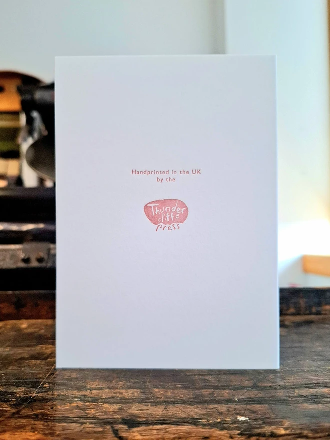 The back of a greetings card standing open on a wooden surface, showing the logo  and text in pink ink.