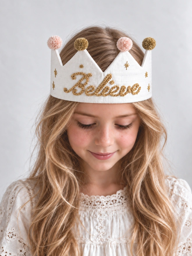 child wearing festive, Christmas "Believe" white silk crown with gold embroidery.