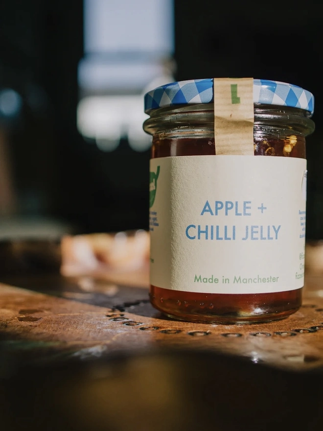 Jar Of Apple & Chilli Jelly, jar of jelly with a blue lid on a decorated plate on a table. 