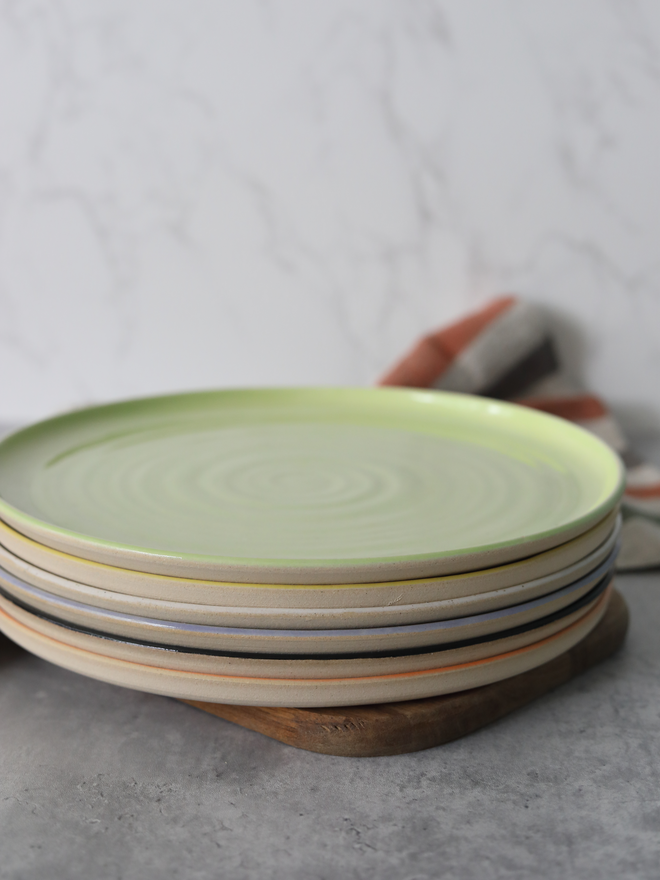A close-up stack of handmade ceramic dinner plates in muted earth tones, topped with a soft lime green plate showing a natural swirl texture, resting on a wooden board against a marble background with a striped linen cloth
