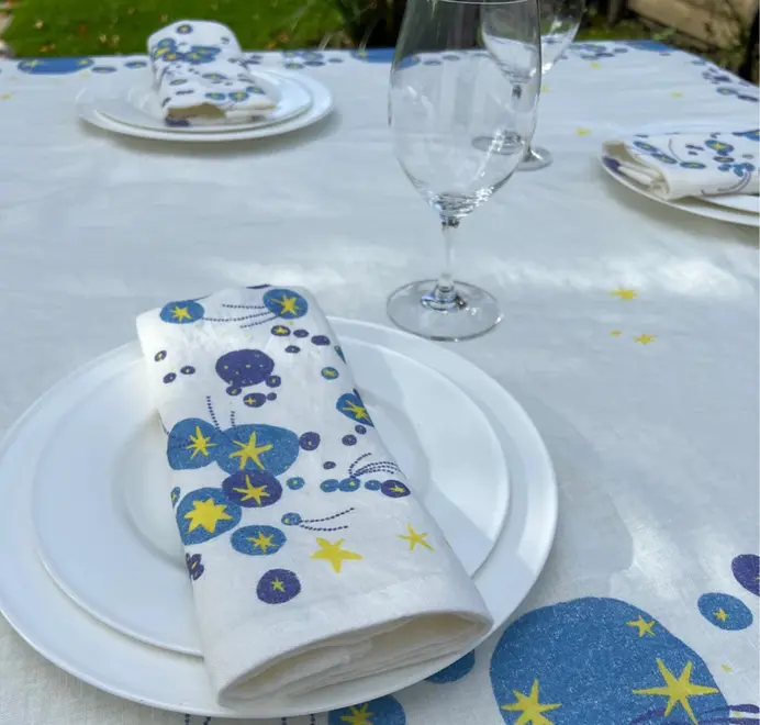 Solaris Napkins In White Or Blue. Off-white cloth napkin with blue and yellow starburst and orbital patterns placed on a plate. 