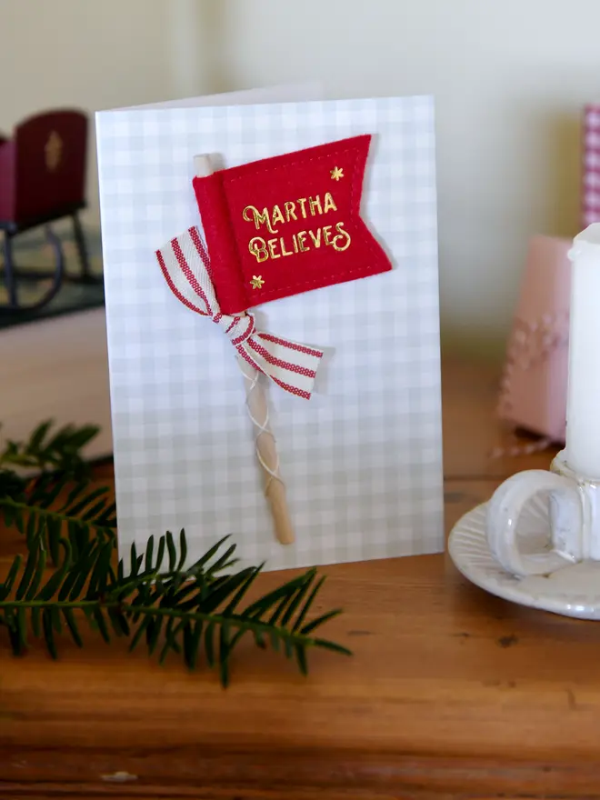 A Christmas card with a tiny handmade felt flag attached. The flag reads 'Martha Believes' and has a red striped ribbon tied to it.