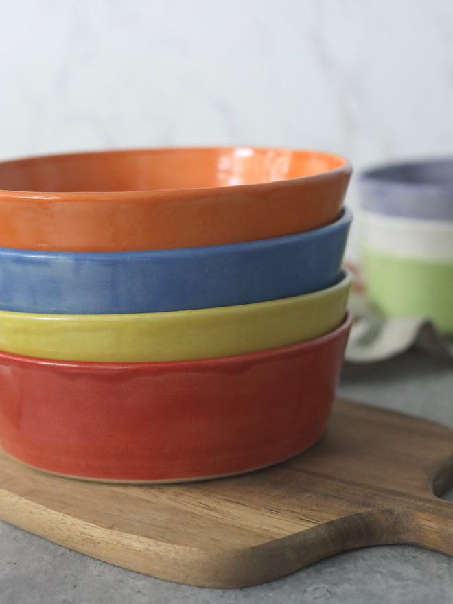 A stack of four glazed ceramic pasta bowls in orange, blue, yellow, and red, resting on a wooden chopping board, with a stack of further pasta bowls partially visible in the background.