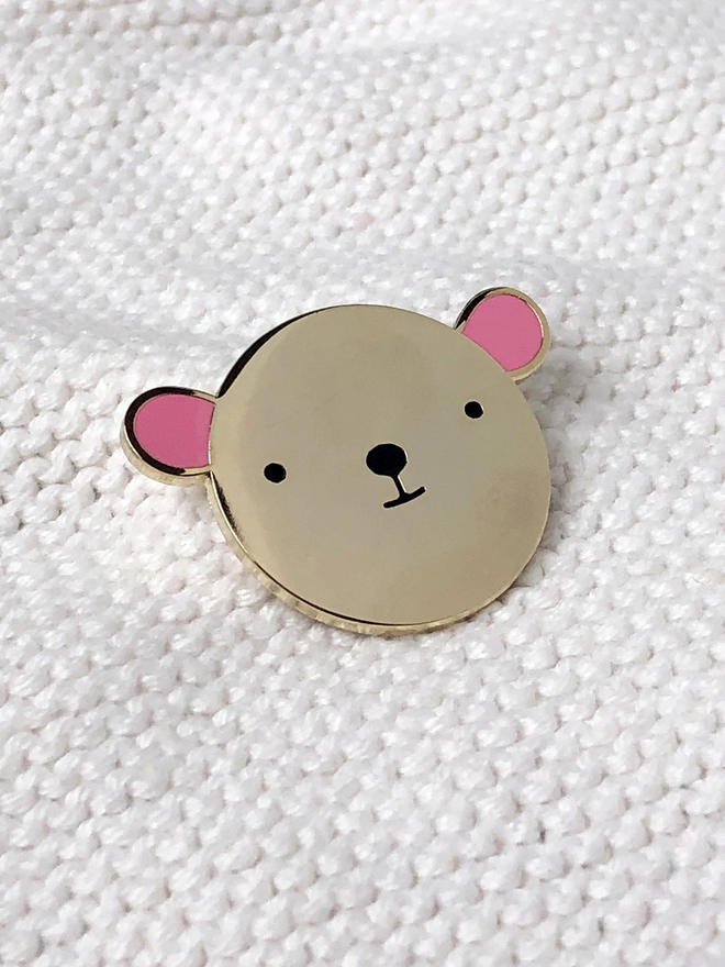 A gold bear with pink ears enamel pin is resting on white fabric.