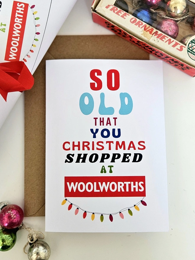 A photograph of the front of a humorous A6 greeting card illustrated using brightly coloured text and the retro shop logo. The text says so old you Christmas shopped at Woolworths with string lights