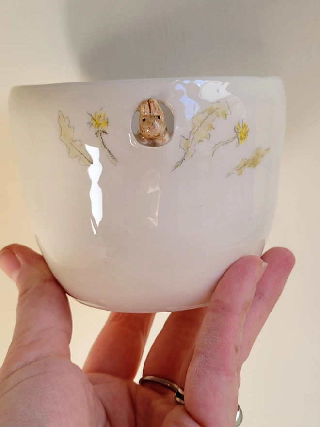 a bunny rabbit cup held in a hand with a bunny looking out of a small cut out and hand painted yellow flowers and green leaves