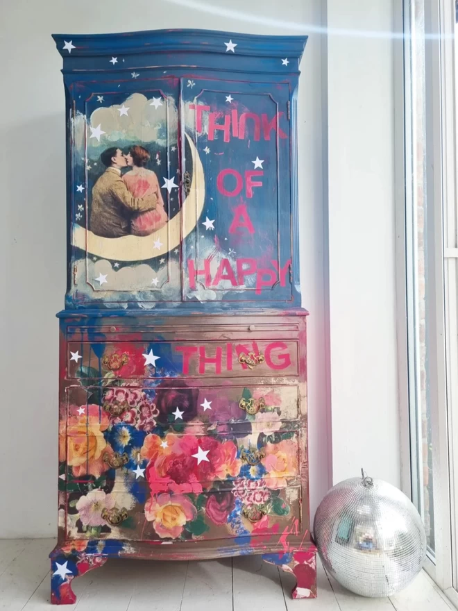 Painted cocktail cabinet featuring flowers and a pair of lovers sitting on a paper moon. The cabinet is painted deep blue and pink and has text that says ‘think of a happy thing’. Quirky and whimsical.