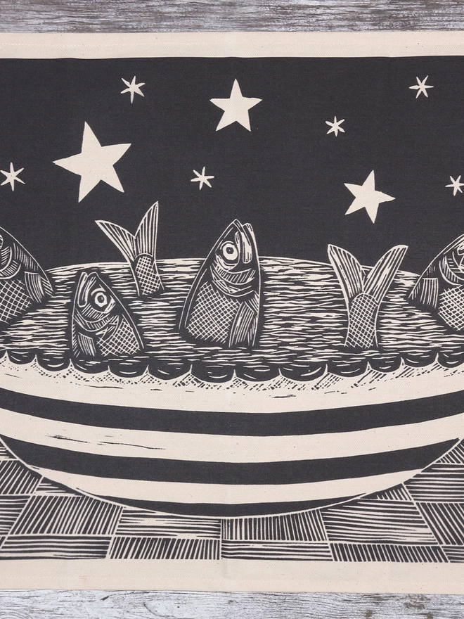 Picture of a tea towel with an image of a fish pie with the fishes gazing up at the stars, taken from an original lino print