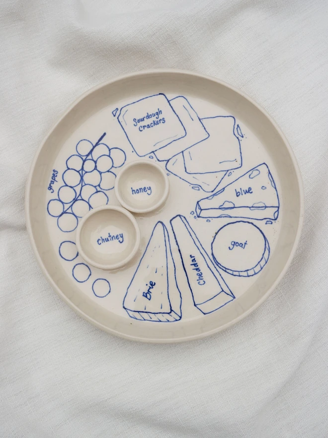 Dippy Cheeseboard Dinner Plate, a white dip plate sitting on a bed cloth. It's covered in blue outlines of cheeses.