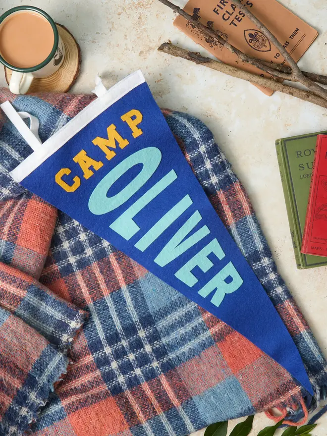Custom camping pennant flag in blue, white and yellow laying with a camping blanket and vintage books