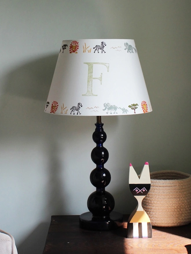 Safari lampshade with letter F, styled on side table in bright boys bedroom. Handmade safari lampshade for kids bedroom, personalised with letter F. Colourful empire light shade with safari animals, a green lampshade option for boys lampshade or kids bedroom design. The lamp is on a dark blue lamp base next to a cream arm chair with blue cushion and sheep skin. 