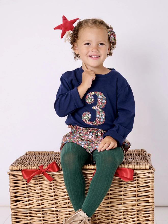 a 3 year old girl on her birthday wearing a navy t-shirt with the number 3 in Liberty print on the front