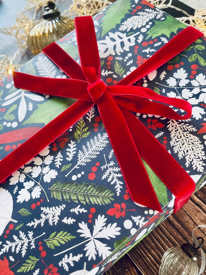 Gift wrapped in festive leaf wrapping paper with Holly and Ivy design, red bow, surrounded by star lights and baubles on wood background