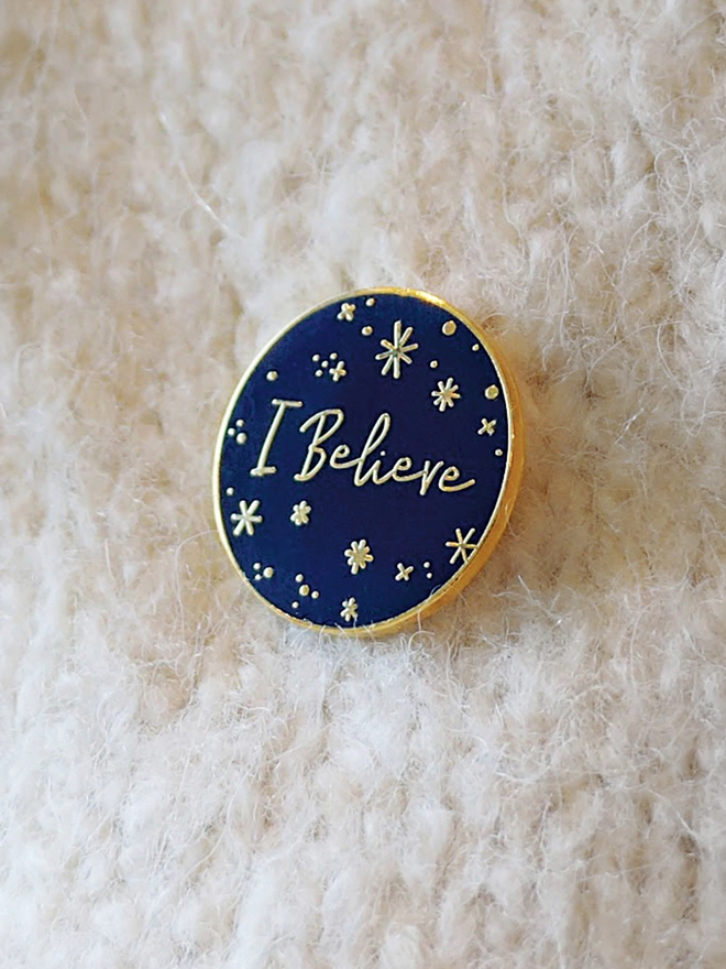 A navy blue and gold enamel pin badge with a starry design and the words "I Believe" is pinned onto a white scarf.