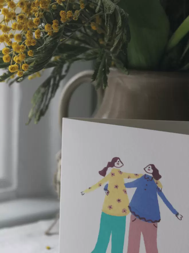 2 girls hugging on a card in front of fresh mimosa flowers 