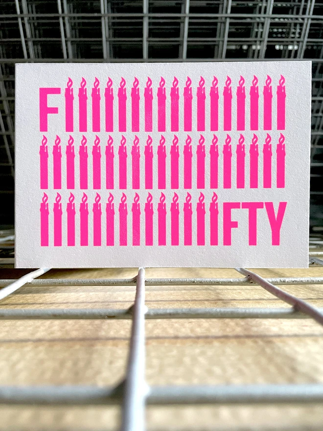 The word Fifty screen printed in neon pink ink across a rectangular recycled white card. To emphasise the Fifty, the letter i is shaped like a simple lit candle and is repeated fifty times in three rows. The card is stood square on, on a drying rack.