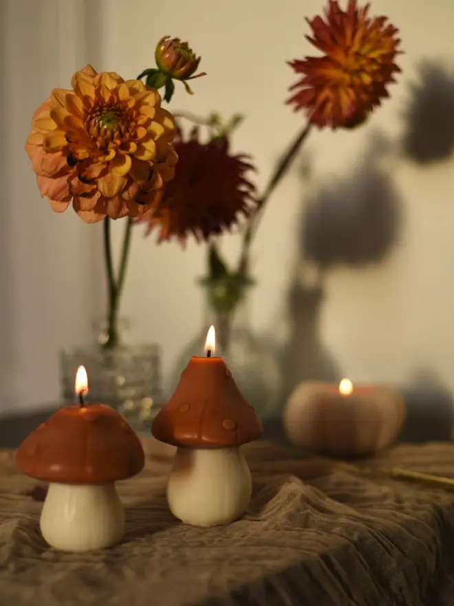 Pair Of Little Mushroom Candles (Set Of 2). Two lit terracotta mushroom candles beside a glowing pumpkin candle and fresh dahlias, set on a crinkled linen tablecloth. The flickering scene creates a warm, inviting mood for autumn evenings and harvest gatherings.