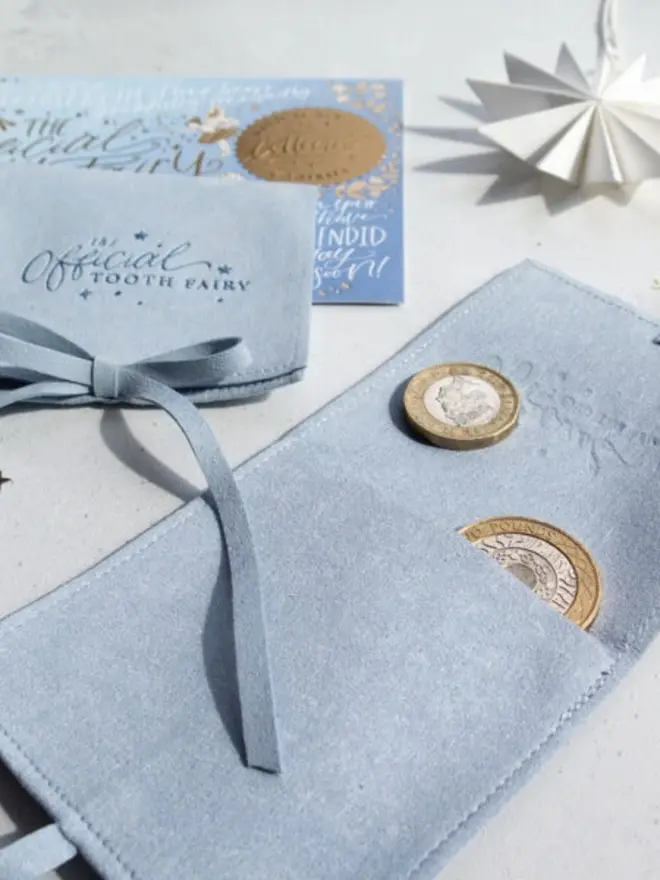 An open pale blue suede pouch holds a pound coin and a two pound coin in its pocket. A second tied pouch and a blue tooth fairy card sit behind it.