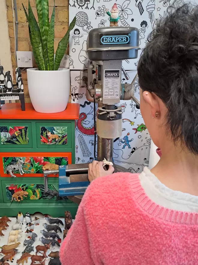 A hand holds a little plastic lamb on a clamp on the table of a pillar drill. A white tray of plastic animals sits below the pillar drill to the left. There is a small chest of drawers decorated in a brightly coloured jungle theme with animal drawer knobs attached with a snake plant in a white vase sitting on top. The lamb is being drilled and made into an animal light switch by Candy Queen Designs.