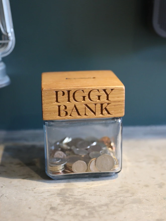 Oak money jar engraved PIGGY BANK, a small glass jar half full of coins.