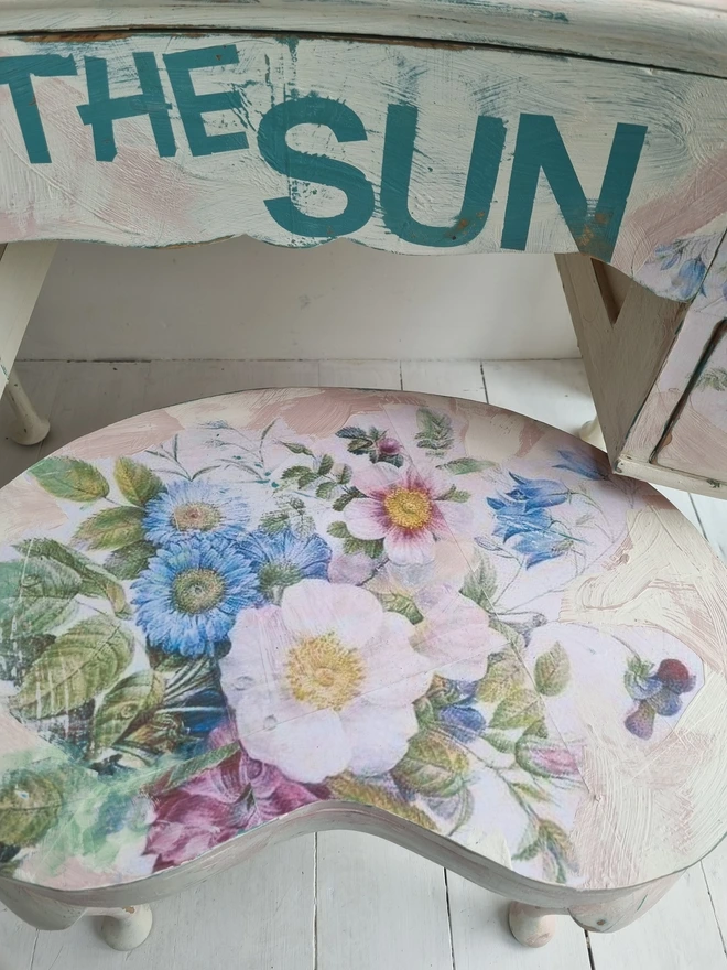 A painted dressing table in white and pale pink with large florals in blues and mauves and text in turquoise that reads 'turn your face to the sun'. The whole piece is distressed in the shabby chic style.