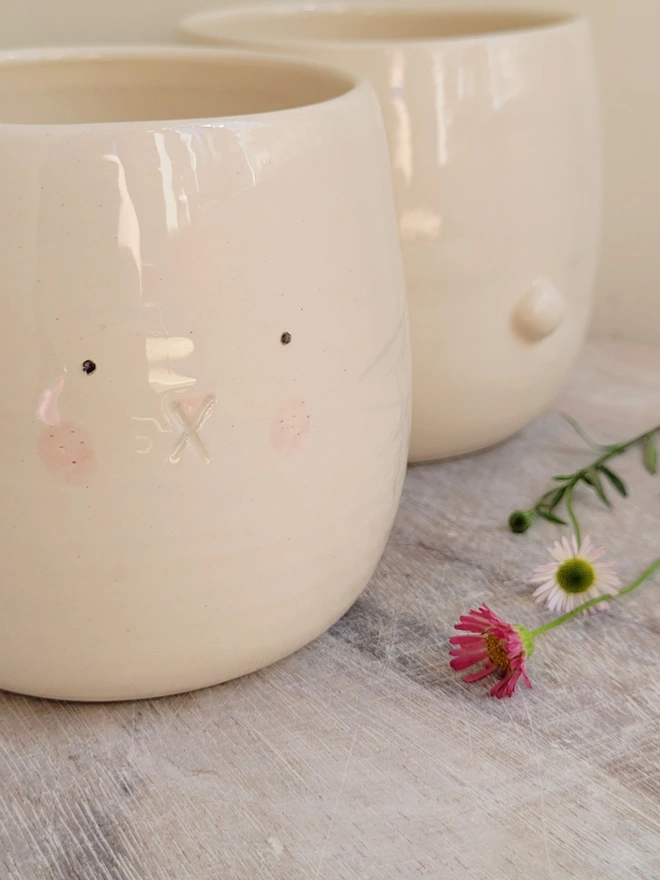 2 pottery cups with a bunny face and bunny tale on a wooden surface with daisies