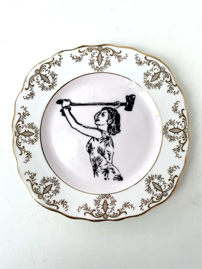A vintage plate with a patterned border , in the centre of the plate is a black and white vintage illustration of a beautiful lady in a floral dress wielding an axe 
