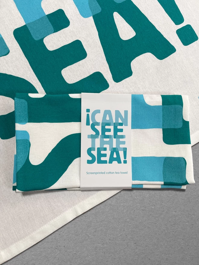 A folded teatowel in a wrap lies on top of an open tea towel, both with the turquoise blue graphic saying I Can See the Sea. On a grey background.