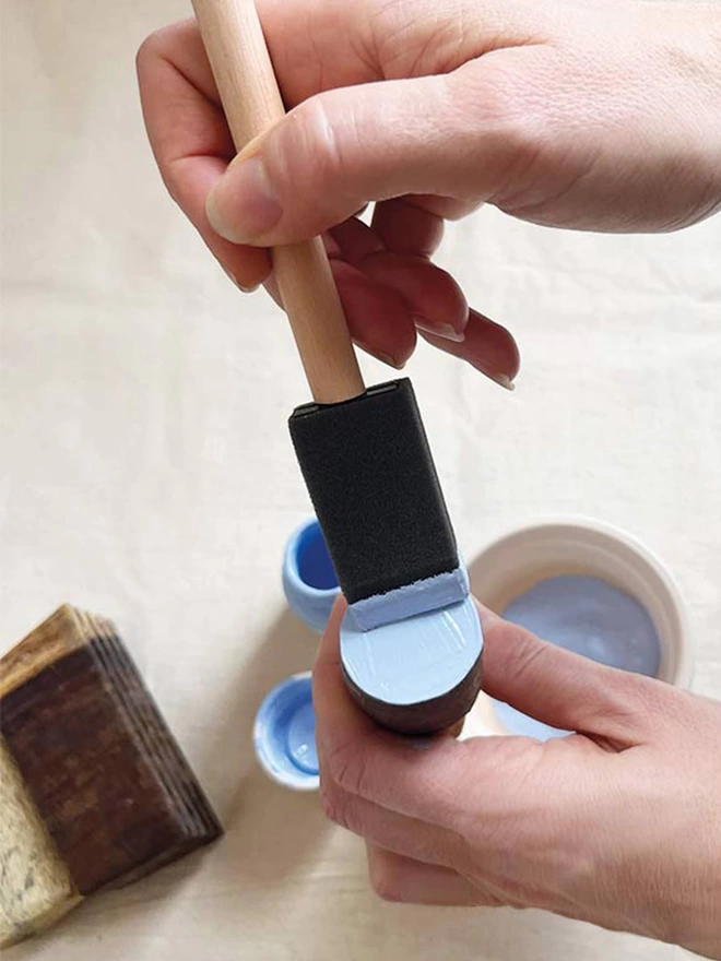 Detail of two hands holding a small circle wooden block and applying pale blue paint with a foam brush