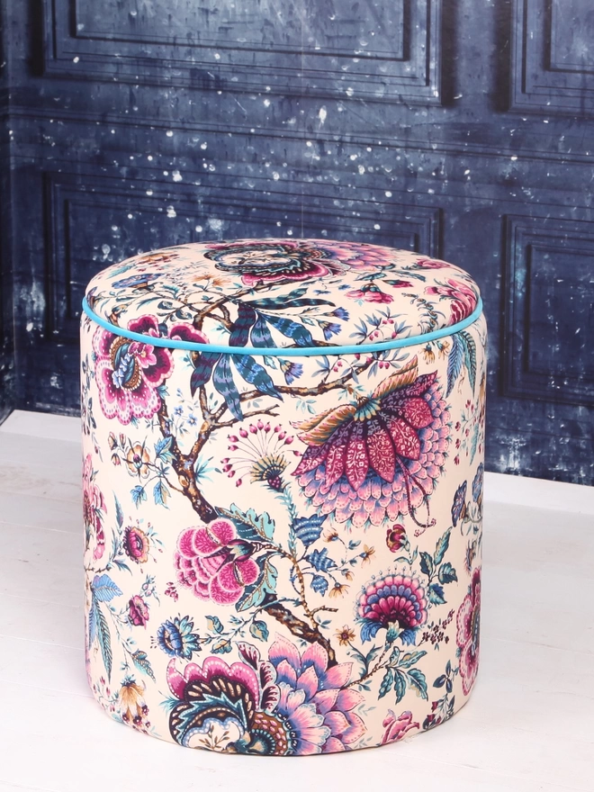 A round pouffe upholstered in a cream and purple floral velvet with tropical blue piping against a blue backdrop
