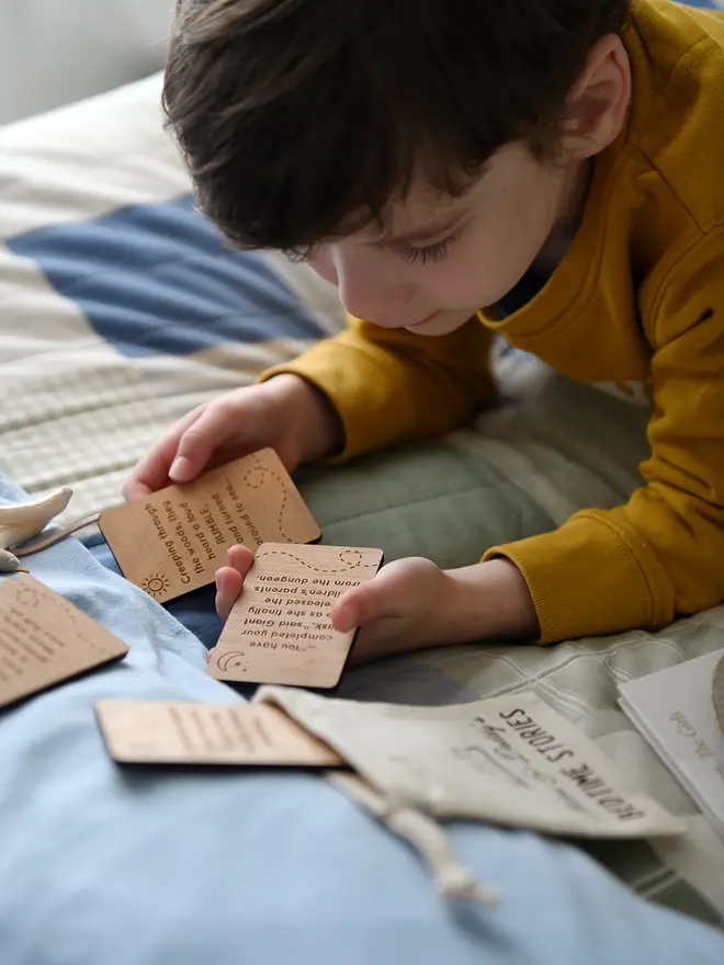 A young child is lying on a bed, covered with a patchwork quilt, reading wooden story prompt cards from a personalised 'Bedtime Stories' bag, designed for storytelling with Daddy.