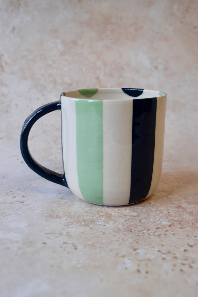Handmade green and cream striped ceramic mug with dark green handle, photographed on a neutral stone background.