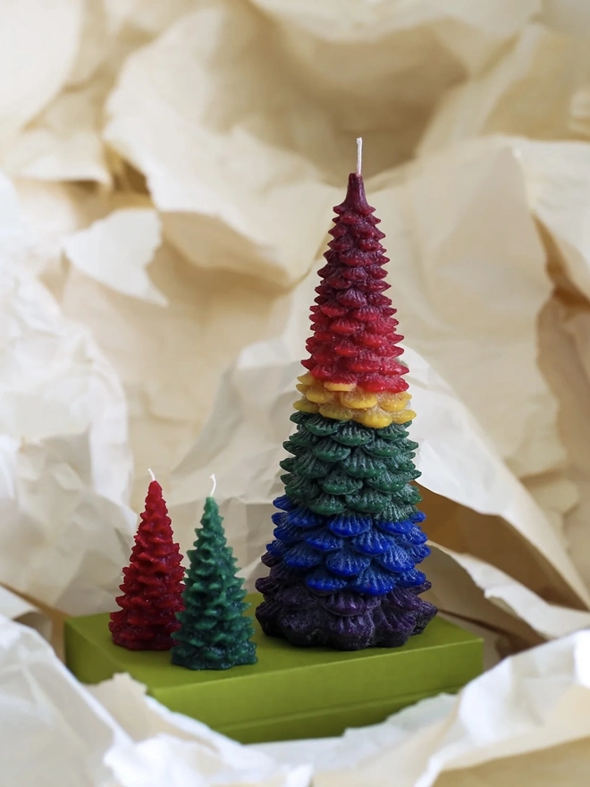 Large Christmas Tree Candle. a large christmas tree shaped candle in a rainbow ombré design of red, yellow, green, blue and purple stands on a green box, nestled among crumpled cream tissue paper. a smaller red and a smaller dark green tree candle sit in front of it for scale, both with visible wicks. the textured, layered pine branch design and multicoloured wax give the large candle a bold, distinctive finish.