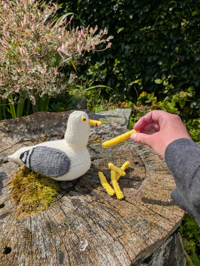 a knitted herring gull being fed knitted chips on a tree stump