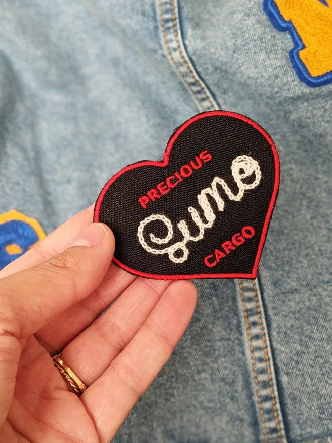 Black & Red 'Precious Cargo' Woven Patch, with Red border on a Black background. The words 'Precious' & 'Cargo' are both woven small above & below the custom chainstitched lettering - reading 'Sumo' & held by hand against a denim jacket that is covered in patches.