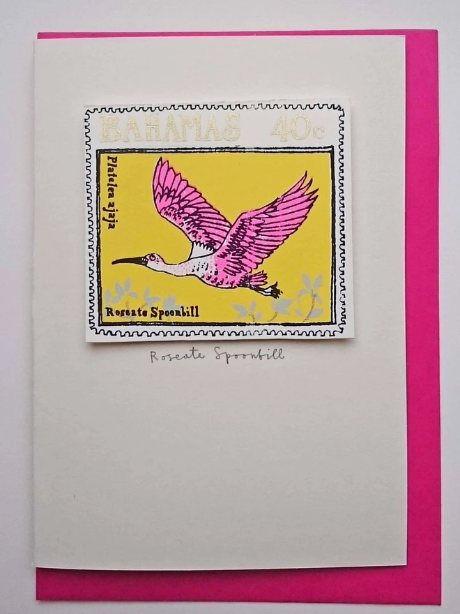 Bird Stamp Card Roseate Spoonbill/ Bahamas, a white card with a colourful bird design in the centre on top of a white surface on a pink envelope. 