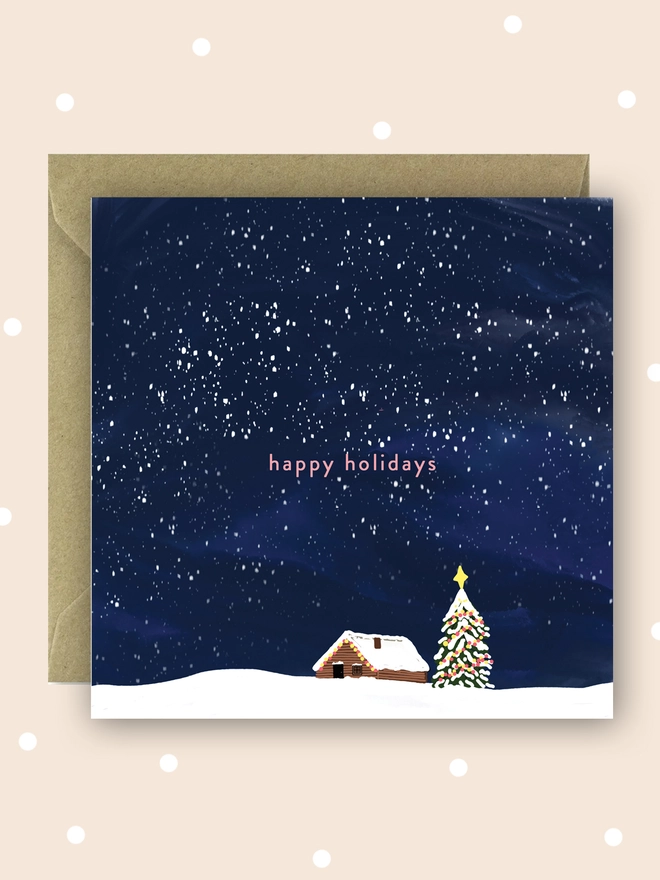 Happy holidays Christmas card with a snowy cabin and Christmas tree under the snowy night sky.