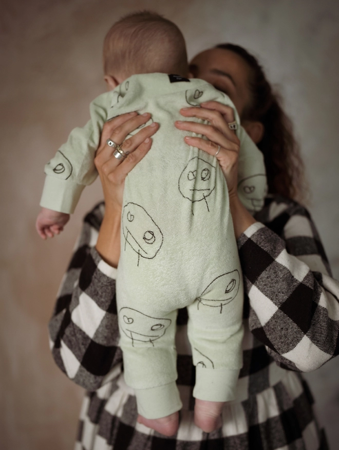 Another Fox Olive Terry Towel Freds Face Baby Sleepsuit seen from behind on a baby held by a woman.