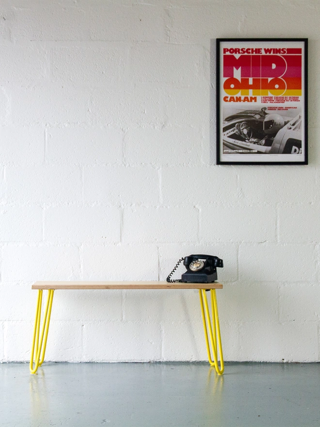 mid century style hairpin leg bench with oak bench top and yellow legs, with an old telephone on it, underneath a classic motorsport poster