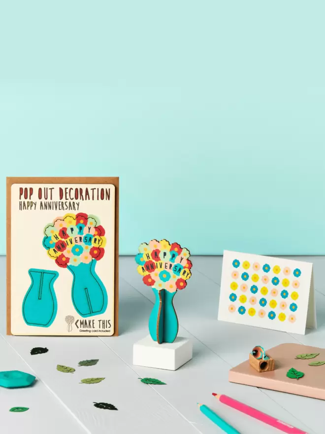 3D vase of flowers happy anniversary decoration and floral pattern anniversary card and brown kraft envelope on top of a wooden desk in front of a pale blue coloured background