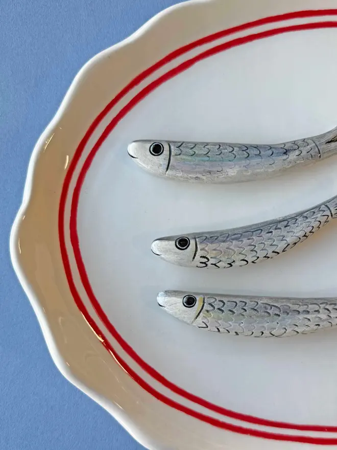 ceramic white platter with red border and anchor detail, with 3 ceramic sardines arranged on top, a ceramic wedge of lemon plus lemon painted on, with parsley detail, on a blue background