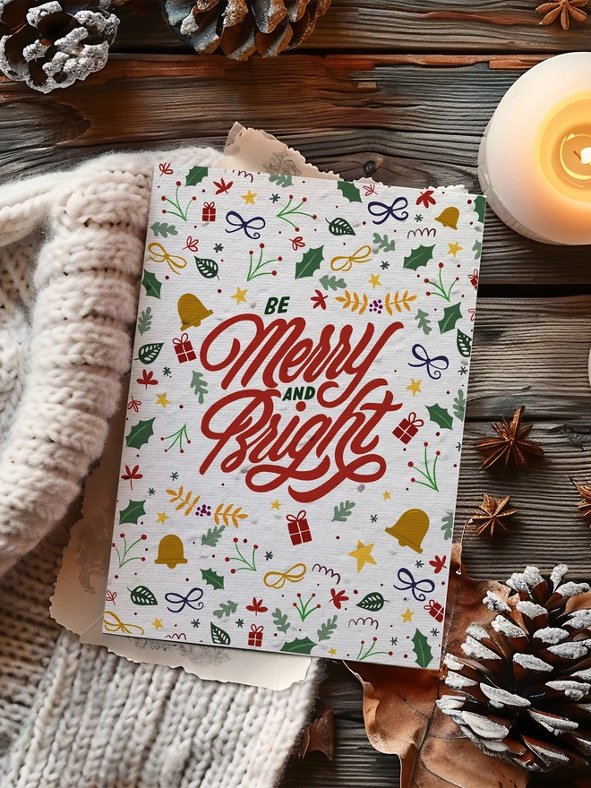 A Christmas card with Merry and Bright in the centre in red with illustrated motifs surrounding it. The card is placed on a wood background with a lit candle, pine cones and cosy blanket.