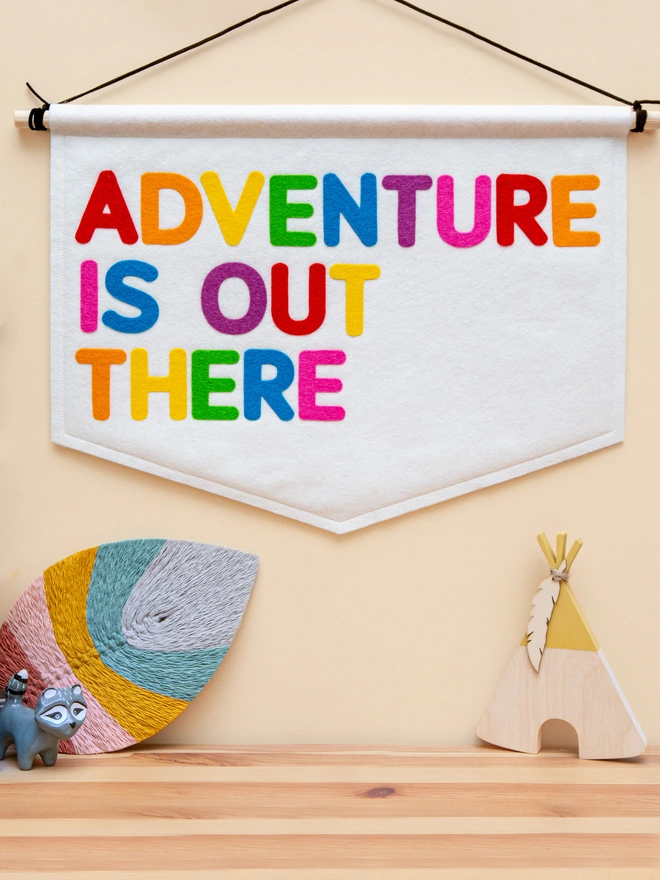 White felt banner with the words "Adventure is out there" written on it in a mix a bright, rainbow colours colours