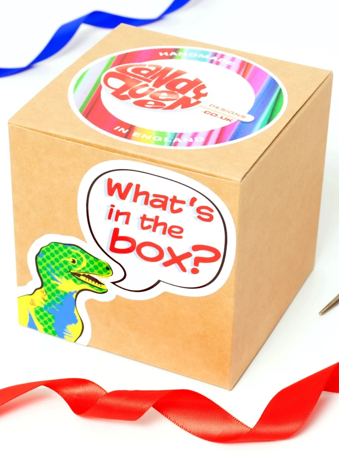 A kraft cardboard box measuring 10 cm x 10 cm x 10 cm. The box has a round sticker on the top with a brightly coloured ring of seaside rock and a lollipop logo for Candy Queen Designs. On the front of the box there is a cartoon style dinosaurs head with a speech bubble that says What's in the Box? There is a string of red ribbon in the foreground and a string of blue ribbon in the background. 