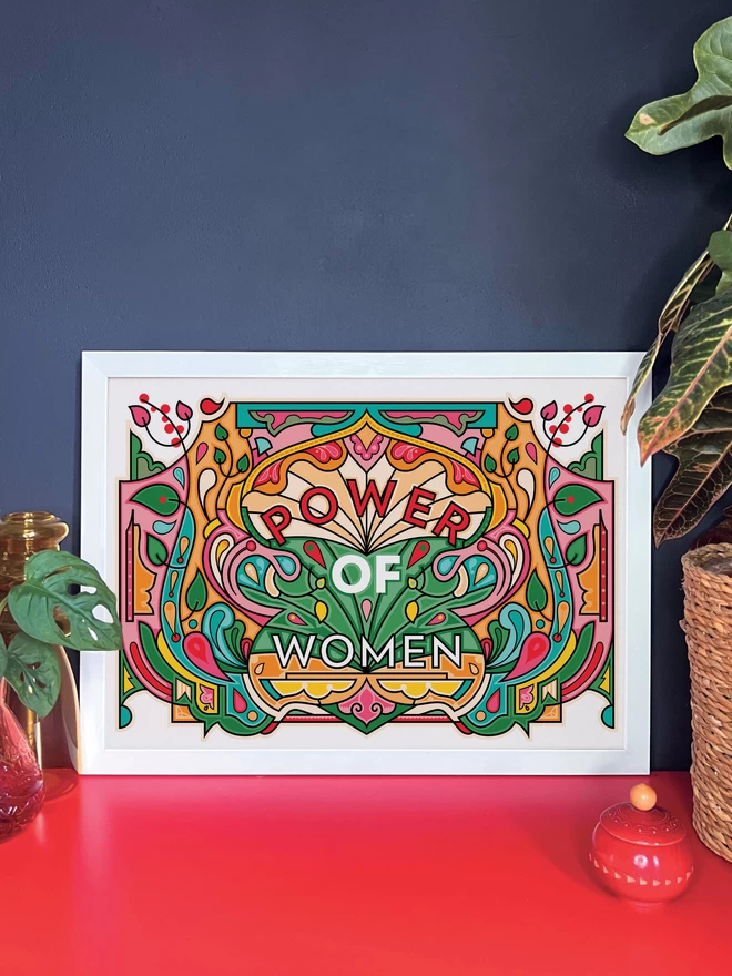 A vibrant, landscape illustration in greens, oranges and reds, with Power of Women written in the centre. The print is in a white frame resting against a dark grey wall on a red cabinet. Next to the frame are two pot plants and a small red wooden pot.