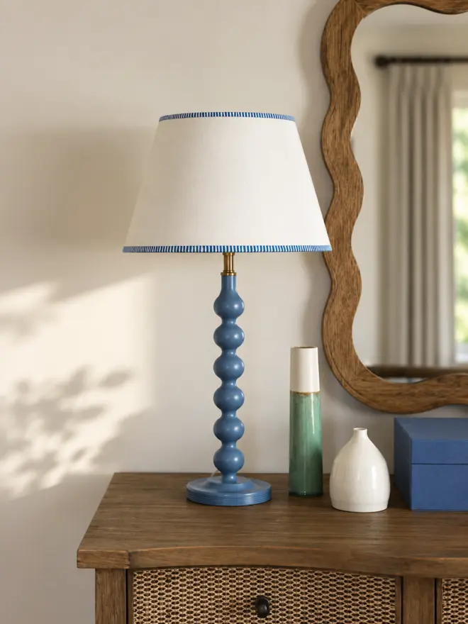 Cream empire lampshade with blue striped trim on a sculptural blue table lamp, styled on a wooden console with ceramic vases and a wavy wooden mirror in a bright contemporary interior.