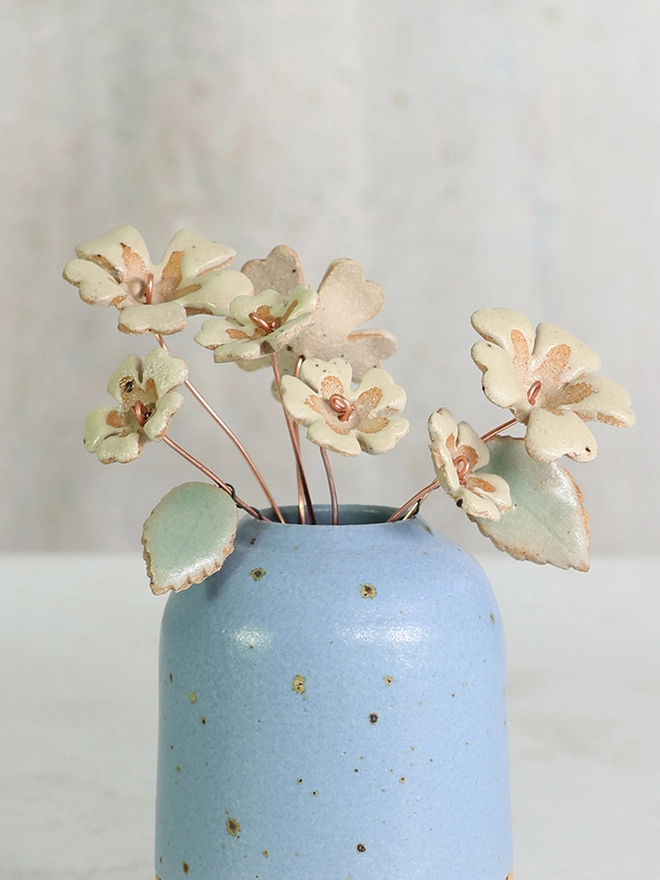 yellow ceramic primrose flowers are arranged in a small blue stoneware vase against a moody grey and white background