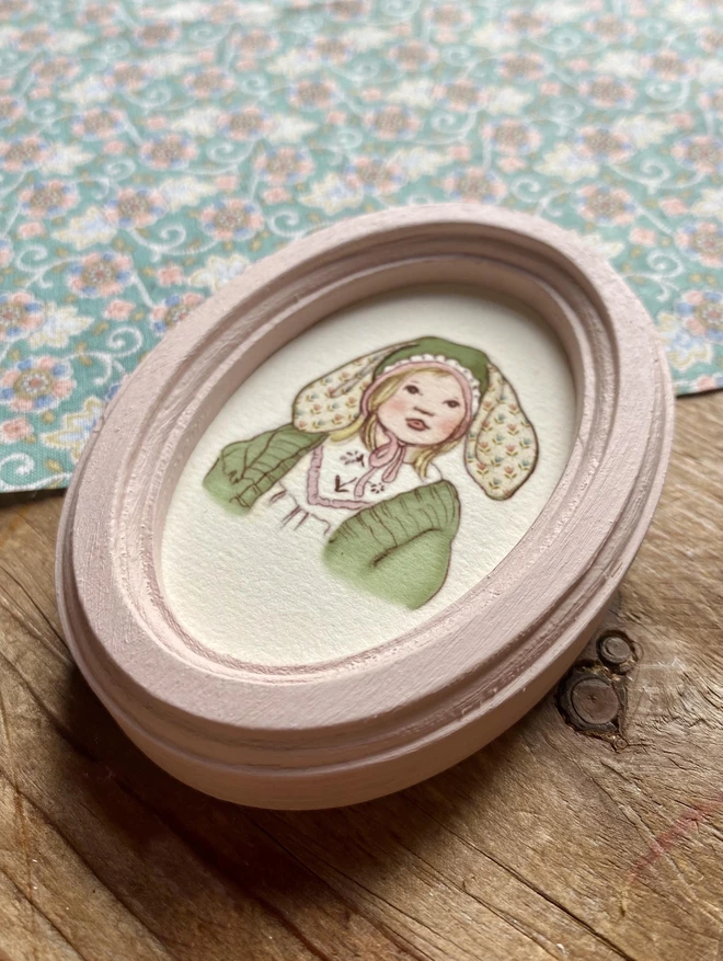 a photograph of a miniature personalised easter bonnet illustration framed in a handmade wooden oval frame