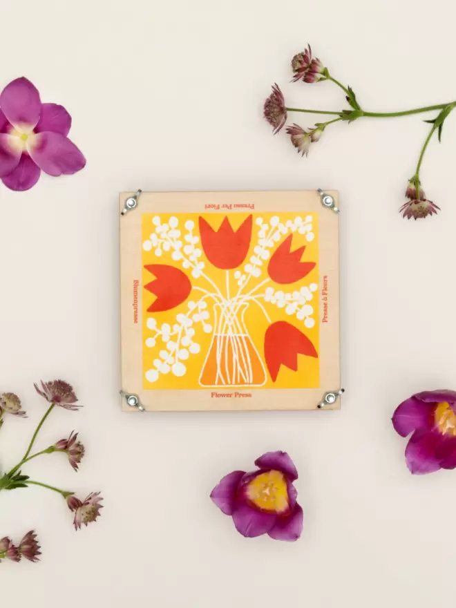 Flatlay of a square flower press with colourful floral front design, surrounded by flowers.