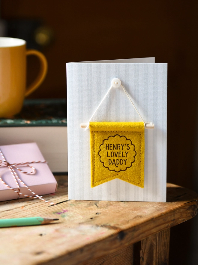 Handmade Father’s Day card with personalised mustard yellow felt banner “Henry's Lovely Daddy”, miniature hanging flag keepsake on wooden table. The flag is hung on the card from a tiny mother of pearl button. Beside the card is a book, a mug, a wrapped gift.